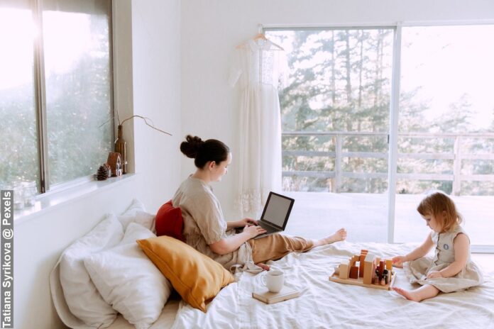 Adorable toddler girl playing with wooden blocks sitting on bed while mother using laptop on sunny morning