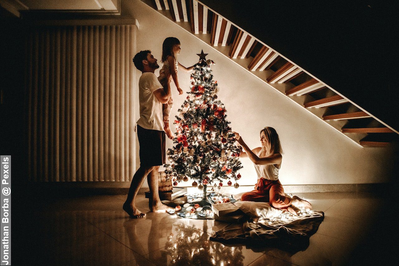 Family decorating their Christmas tree, mum, dad and daughter in the dark under stairs with Christmas tree lights on