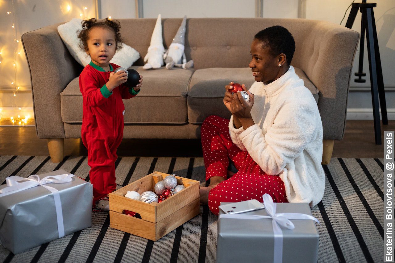 Little Mixed race girl standing holding Christmas tree balls and mother sitting on the floor watching and helping her to decorate