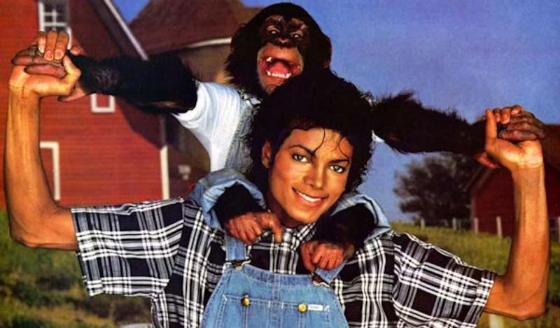 Michael Jackson smiling in a plaid shirt and denim overalls with his pet chimpanzee Bubbles perched on his shoulders, holding his hands, in front of a rural barn backdrop.